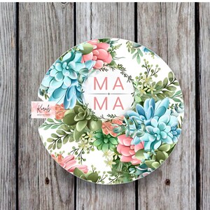 May include: A white round coaster with a floral design featuring pink and blue succulents. The center of the coaster has a wreath with the word "MAMA" in pink letters.