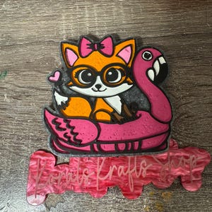 May include: A whimsical, orange fox wearing glasses and a pink bow, floating on a pink flamingo floatie. The fox is holding a wooden stick. The image has a pink and red sign that says "Kora's Krafts Shop".