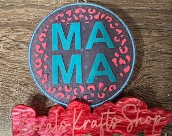 May include: A circular purple ornament featuring a pink leopard print pattern and the word MAMA in large teal letters. Attached below is a red, marbled, cloud-shaped plaque with the text Korals Krafts Shop in a cursive, light pink font.