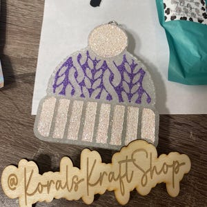 May include: A white and purple glitter winter hat ornament with a white pom-pom on top. The ornament is made of resin and has a glittery finish. The ornament is decorated with a purple glitter design. The ornament is hanging from a silver string. The ornament is on a wooden surface with the text "@Korals Kraft Shop" written on it.