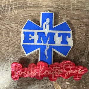 May include: A blue and white star of life with the word "EMT" in white letters. The star of life is a symbol used by emergency medical technicians. The star of life is on a wooden surface with a red sign that says "Korals Krafts Shop".