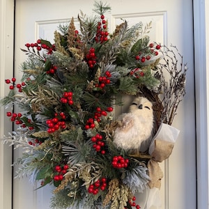 May include: A festive wreath with a plush owl, red berries, and faux greenery. The wreath is adorned with pine needles, silver leaves, and a burlap bow. This decorative wreath is perfect for the holiday season.