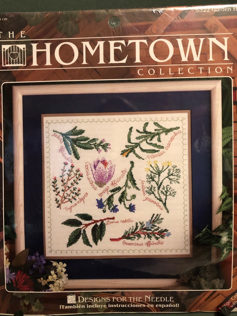 Może przedstawiać: Wzorzec haftu krzyżykowego w ramie z białym tłem i r&oacute;żnymi zielonymi ziołami i kwiatami. Nazwy zi&oacute;ł i kwiat&oacute;w są napisane czarnym kolorem wok&oacute;ł obrazu. Rama jest wykonana z jasnego brązowego drewna z ciemnoniebieskim passe-partout.