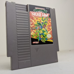 May include: A gray Nintendo Entertainment System (NES) game cartridge with a label featuring the Teenage Mutant Ninja Turtles. The label has a yellow background with a black border and the text "Teenage Mutant Ninja Turtles The Arcade Game" in white. The image shows four turtles in action, with a red "Ultra Games" logo in the bottom left corner.