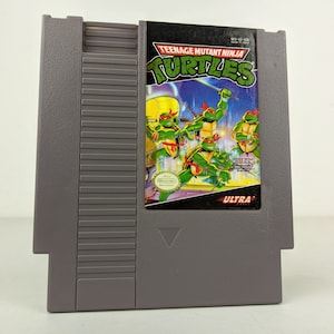 May include: A gray Nintendo Entertainment System (NES) game cartridge for Teenage Mutant Ninja Turtles. The cartridge features a colorful image of the four Teenage Mutant Ninja Turtles fighting on a futuristic cityscape. The text "Teenage Mutant Ninja Turtles" is printed in white on a black background. The text "Ultra" is printed in white on a red background.