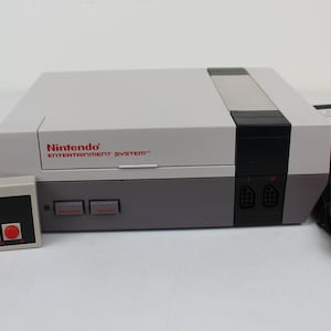 May include: A Nintendo Entertainment System (NES) console with a controller and power supply. The console is gray and dark gray with red lettering. The controller is gray with a black directional pad and red buttons.