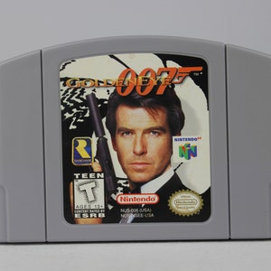 May include: A grey Nintendo 64 game cartridge with a black label featuring a photo of Pierce Brosnan as James Bond from the film GoldenEye. The label also includes the game title "GoldenEye 007", the Nintendo 64 logo, and the ESRB rating "Teen".