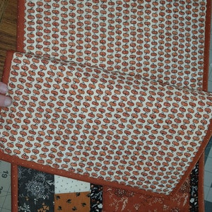 May include: A set of two quilted placemats featuring a patchwork design with a brown, black, and white floral pattern. The placemats are bordered with a solid brown fabric. One placemat has a pumpkin pattern.