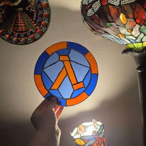 Puede incluir: Un medallón de vidriera con un símbolo lambda azul y naranja, sostenido en la mano. El medallón tiene un borde azul y naranja. Otras lámparas de vidriera son visibles en el fondo, incluida una lámpara de libélula.