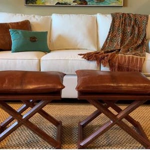 Peut inclure: Scène de salon avec un canapé blanc, des coussins en cuir marron et turquoise, et une couverture à motifs. Deux poufs en cuir marron avec des bases en bois en forme de X sont placés devant le canapé, créant un espace confortable et élégant.