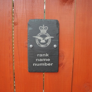 May include: A dark grey slate sign featuring a silver emblem of a crown and bird. The sign is inscribed with the words "rank name number". Two silver rivets attach the sign to a wooden surface. The background is a wooden fence.