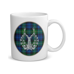 May include: White ceramic mug with a circular design showcasing a blue, green, and black tartan pattern. The design includes a silver stag's head with antlers, a floral wreath, and the word "BYDAND" on a banner.
