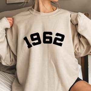May include: A beige sweatshirt with black text that reads "1962" in a bold, blocky font.