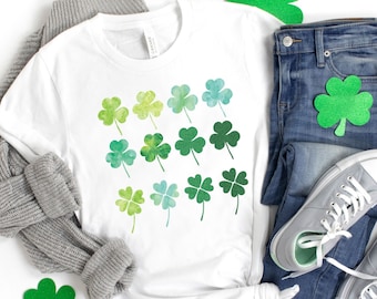 May include: A white t-shirt featuring a green watercolour shamrock design. The shirt is laid out on a white background with a grey sweater, blue jeans, and green shamrock decorations.