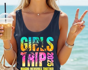 May include: Navy blue tank top with the words "GIRLS TRIP 2025" in a colorful, tropical design. The text "MAKING MEMORIES TOGETHER YOUR DESTINATION" is below the main text. The image shows a person holding a drink and a straw bag.