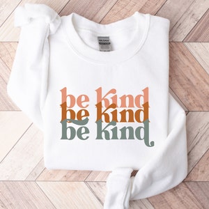May include: White sweatshirt with the phrase "be kind" repeated three times in stacked, overlapping, retro-style font. The text is in shades of peach, orange, and green.