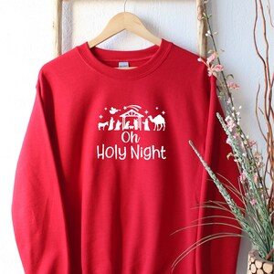 K&ouml;nnte beinhalten: Roter Weihnachtspullover mit einem wei&szlig;en Design einer Weihnachtskrippe und dem Text "Oh Holy Night".