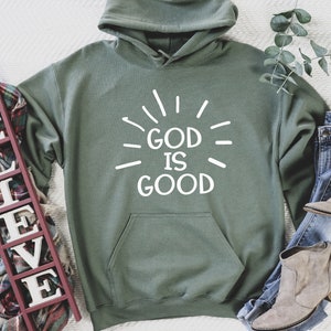May include: A green hooded sweatshirt with the text "God is Good" in white lettering. The sweatshirt is laid flat on a white surface with a wooden sign that says "Believe" and a pair of brown boots.