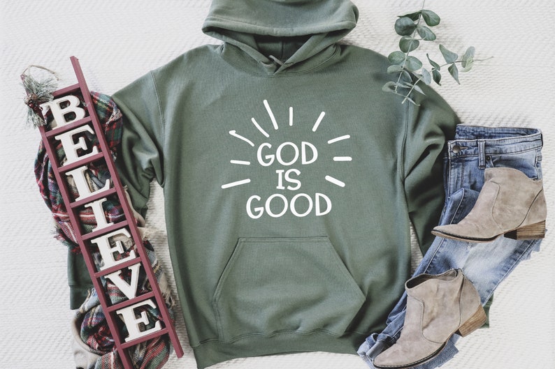 May include: A green hooded sweatshirt with the text "God is Good" in white lettering. The sweatshirt is laid flat on a white surface with a wooden sign that says "Believe" and a pair of brown boots.