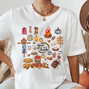 May include: White t-shirt featuring a colorful Thanksgiving-themed graphic. The design includes pumpkins, a scarecrow, a bonfire, and the words "give thanks". Other elements include a turkey, pie, and fall foliage.