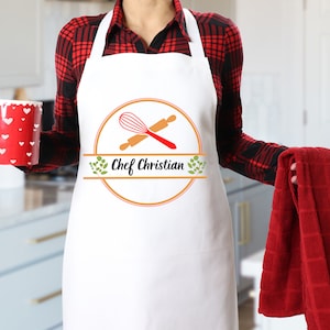 May include: White apron with a red and black checked shirt underneath. The apron has a logo with a whisk and rolling pin crossed over each other, with the text "Chef Christian" in a circle.