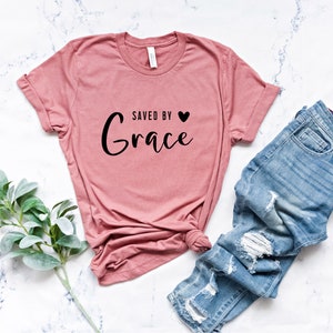 K&ouml;nnte beinhalten: Ein rosa T-Shirt mit dem Text "Gerettet durch Gnade" und einem kleinen schwarzen Herz.