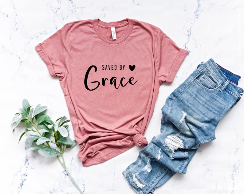 K&ouml;nnte beinhalten: Ein rosa T-Shirt mit dem Text "Gerettet durch Gnade" und einem kleinen schwarzen Herz.