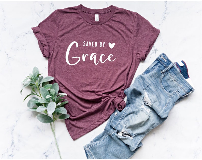 K&ouml;nnte beinhalten: Ein maroonfarbenes T-Shirt mit dem Text "Saved by Grace" in wei&szlig;en Buchstaben mit einem kleinen wei&szlig;en Herz.