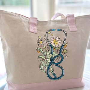 May include: A beige canvas tote bag with pink handles and a pink bottom border. The bag features a blue stethoscope with a floral design in shades of yellow, pink, and green.