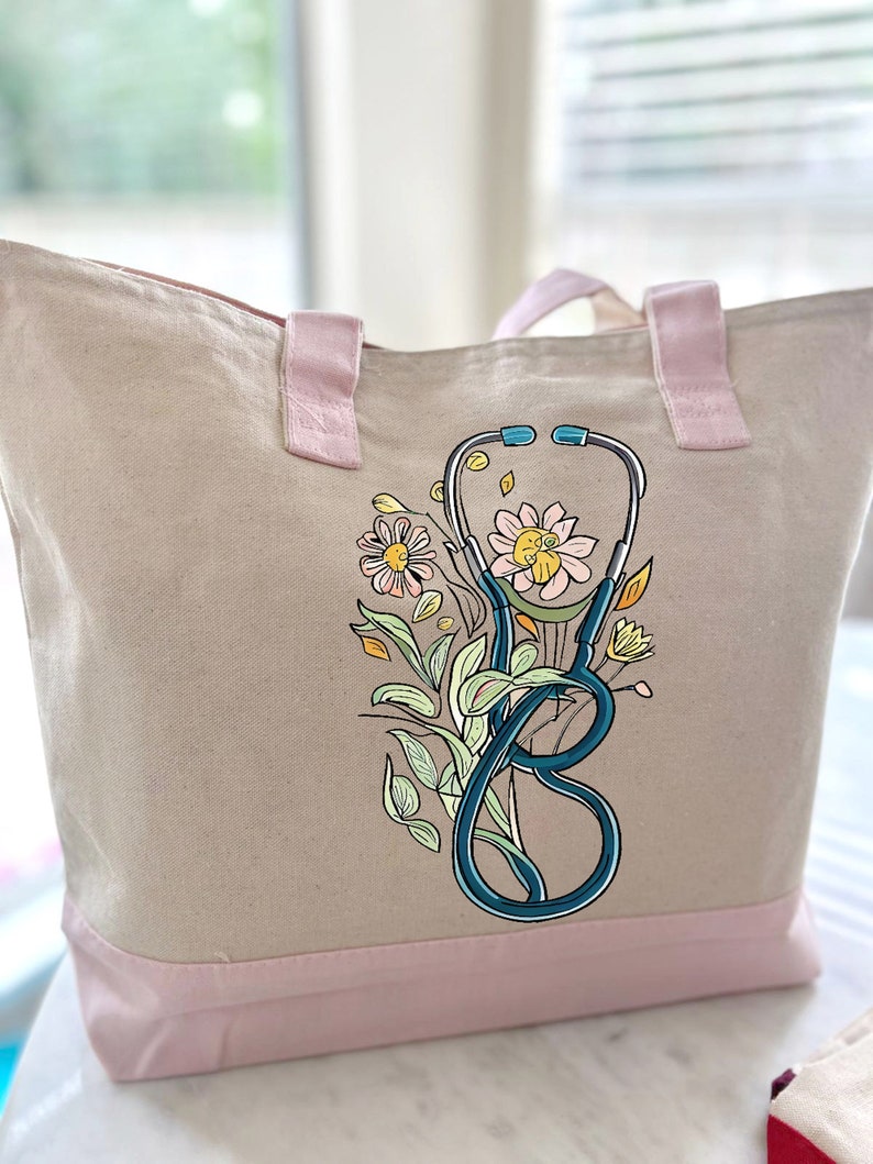 May include: A beige canvas tote bag with pink handles and a pink bottom border. The bag features a blue stethoscope with a floral design in shades of yellow, pink, and green.