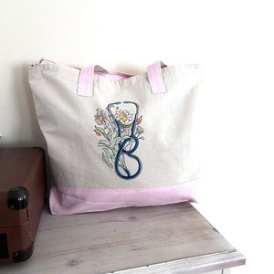 May include: A white canvas tote bag with pink handles and a pink bottom. The bag has a blue and black stethoscope with flowers embroidered on it.