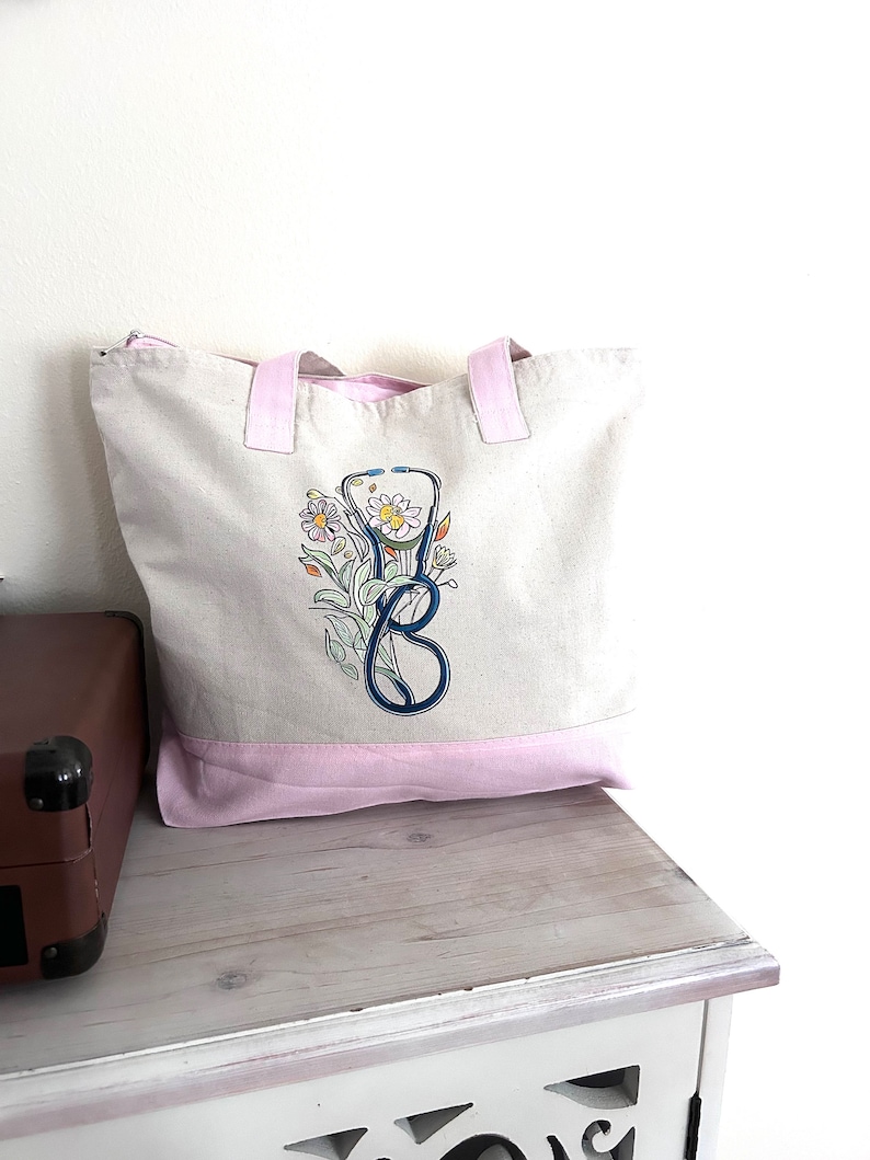 May include: A white canvas tote bag with pink handles and a pink bottom. The bag has a blue and black stethoscope with flowers embroidered on it.