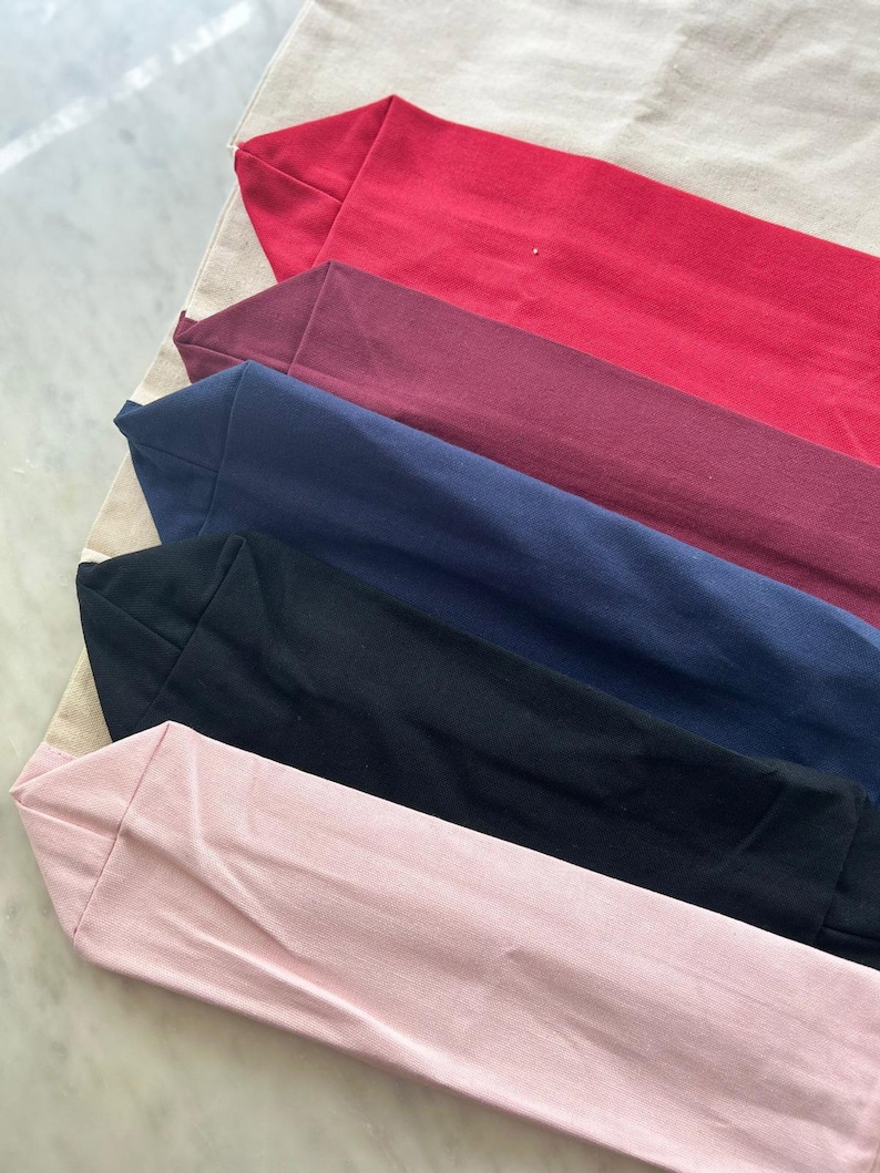 May include: Five tote bags in different colors, including red, burgundy, navy blue, black, and pink. The bags are made of canvas and are folded on top of each other.