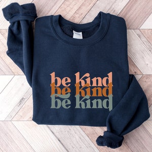 May include: A navy blue sweatshirt with the phrase "be kind" repeated three times in stacked, overlapping, colorful letters. The letters are in shades of orange, brown, and green.