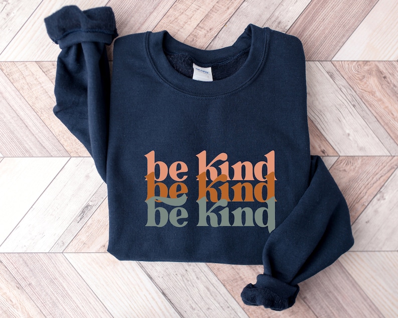 May include: A navy blue sweatshirt with the phrase "be kind" repeated three times in stacked, overlapping, colorful letters. The letters are in shades of orange, brown, and green.