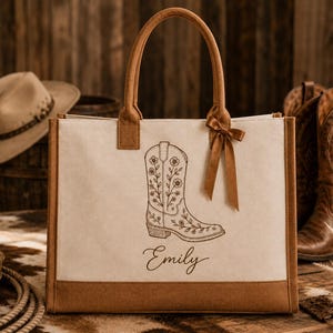 May include: A beige canvas tote bag with brown trim and handles, featuring a brown ribbon bow on the right handle. The front displays a dark brown line art illustration of a floral cowboy boot above the name Emily in a cursive script font.