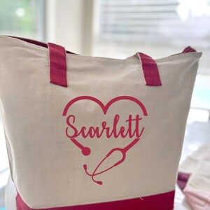 May include: A white canvas tote bag with red handles and a red heart design. The heart has a stethoscope inside and the name "Scarlett" written inside the heart.