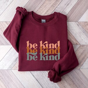 May include: Burgundy sweatshirt with the phrase "be kind" repeated three times in stacked, overlapping, retro-style font. The text is in shades of peach, orange, and green.