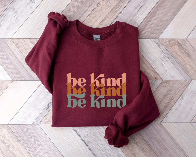 May include: Burgundy sweatshirt with the phrase "be kind" repeated three times in stacked, overlapping, retro-style font. The text is in shades of peach, orange, and green.