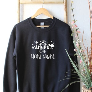 K&ouml;nnte beinhalten: Schwarzer Pullover mit einem wei&szlig;en Aufdruck einer Weihnachtskrippe und dem Text "Oh Holy Night".