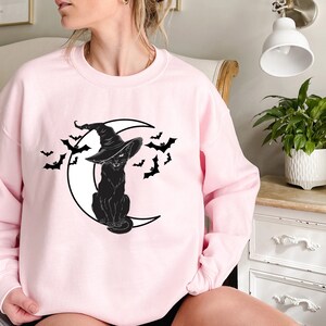 May include: A light pink sweatshirt featuring a black cat wearing a witch hat, sitting on a crescent moon, with bats flying around. The design is a Halloween theme.