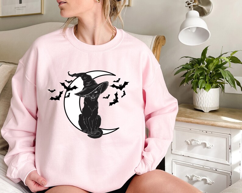 May include: A light pink sweatshirt featuring a black cat wearing a witch hat, sitting on a crescent moon, with bats flying around. The design is a Halloween theme.
