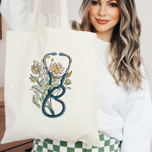 May include: A white canvas tote bag with a blue stethoscope and colorful flowers printed on it. The bag has a long handle and is made of natural fabric.