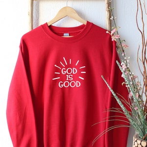 May include: Red sweatshirt with a white graphic that says "God is Good" with radiating lines around the text.