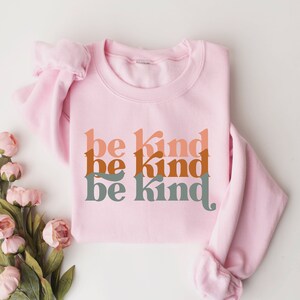 May include: Pink sweatshirt with the phrase "be kind" repeated three times in a stacked design. The text is in a retro font style with shades of orange, brown, and green.