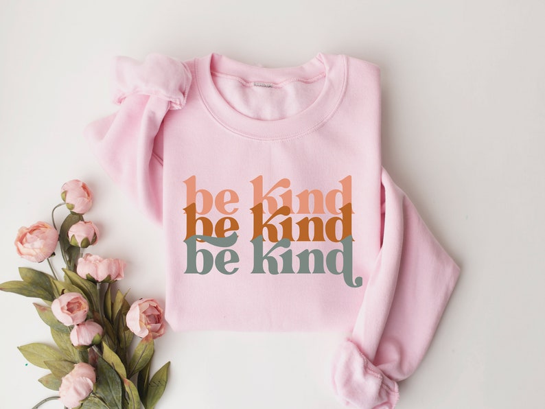May include: Pink sweatshirt with the phrase "be kind" repeated three times in a stacked design. The text is in a retro font style with shades of orange, brown, and green.