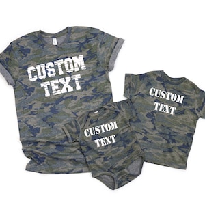 May include: Two short-sleeve t-shirts and one short-sleeve bodysuit in a green, brown, and navy blue camouflage pattern. Each garment features the white text CUSTOM TEXT printed in a bold, distressed font across the centre of the chest area.
