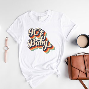 May include: White t-shirt with a retro-style graphic that says "90's Baby" in rainbow colors.