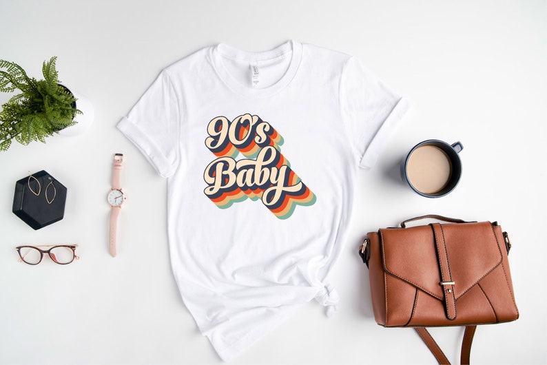 May include: White t-shirt with a retro-style graphic that says "90's Baby" in rainbow colors.