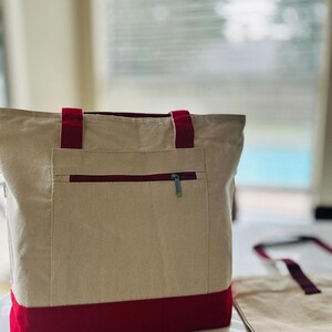 May include: A beige canvas tote bag with a red trim and a zippered pocket. The bag has two red straps.